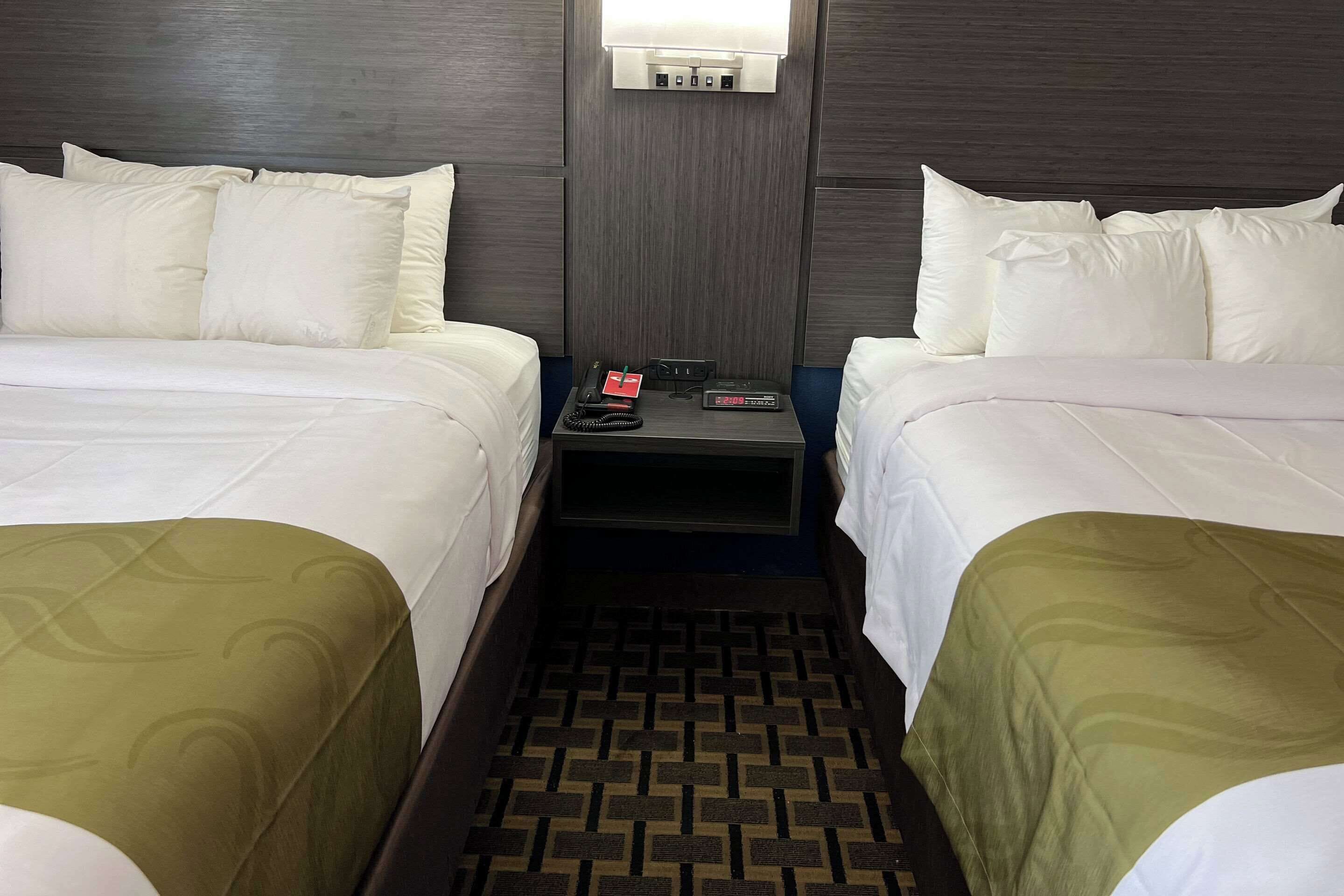 Habitación de hotel con dos camas, una mesita de noche con un teléfono y un control remoto, y un aire acondicionado instalado en la pared.