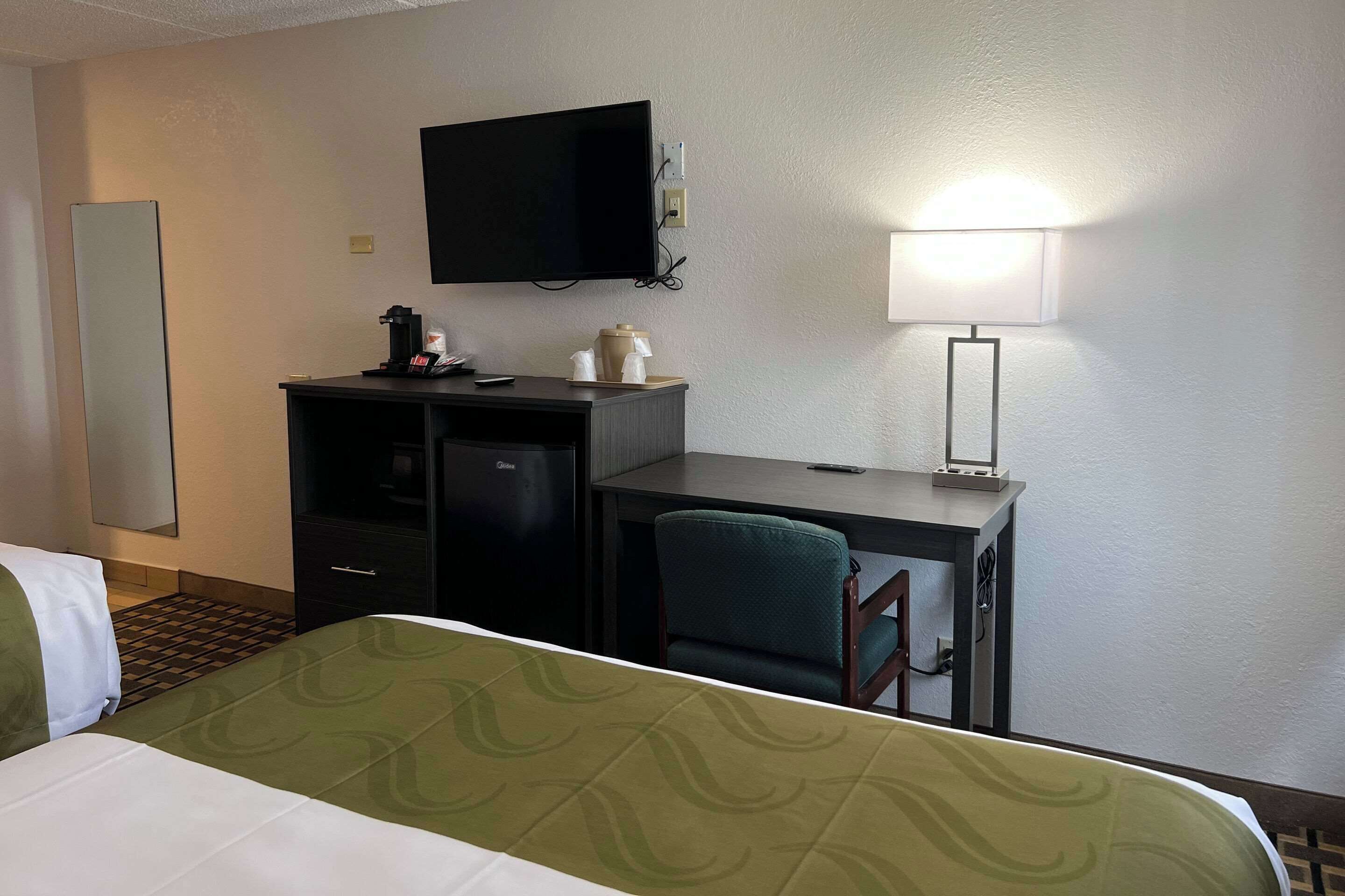 Habitación de hotel con cama, escritorio con silla, televisor, lámpara y minibar.