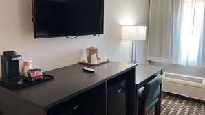 Une chambre d’hôtel équipée d’un téléviseur à écran plat, d’une machine à café, d’une télécommande, d’un petit réfrigérateur, d’un bureau, d’une chaise, d’une lampe et d’une fenêtre avec des rideaux.