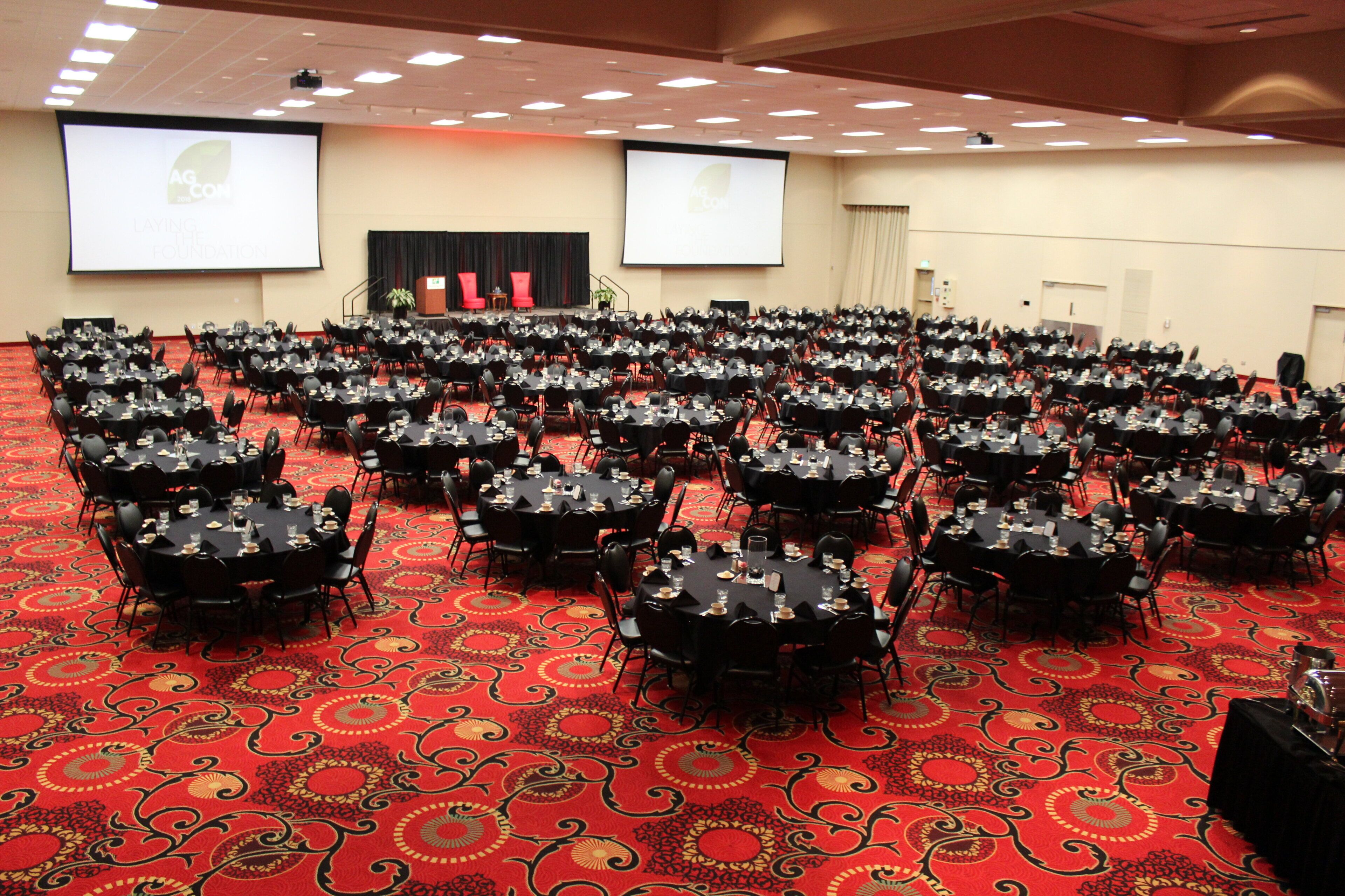 Une grande salle de conférence, dotée de nombreuses tables et chaises disposées en rangées, avec un sol recouvert de moquette et une estrade à l’avant.