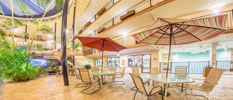 A spacious indoor area with multiple levels, featuring a central seating area with tables and chairs, surrounded by balconies with plants.