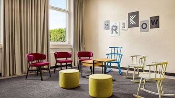 Une pièce avec une grande fenêtre, des rideaux beiges, une moquette beige et une table avec des chaises. Des lettres encadrées sont accrochées au mur.
