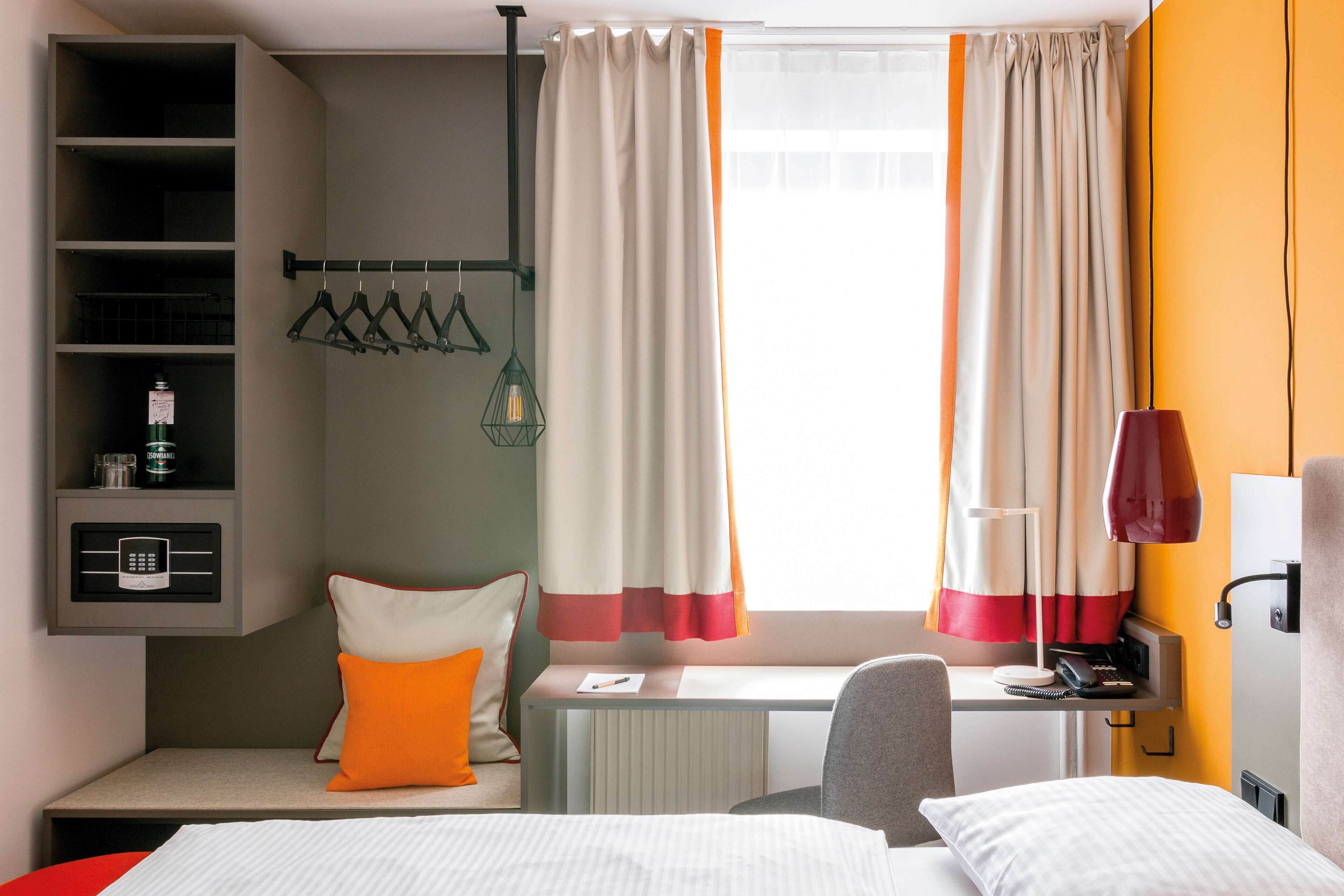 Habitación de hotel con cama, escritorio, silla, radiador, estante con microondas, lámpara colgante y ventana con cortinas.