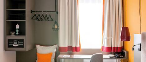 Habitación de hotel con cama, escritorio, silla, radiador, estante con microondas, lámpara colgante y ventana con cortinas.