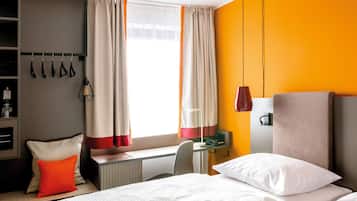Habitación de hotel con cama, escritorio, silla, radiador, armario y ventana con cortinas.