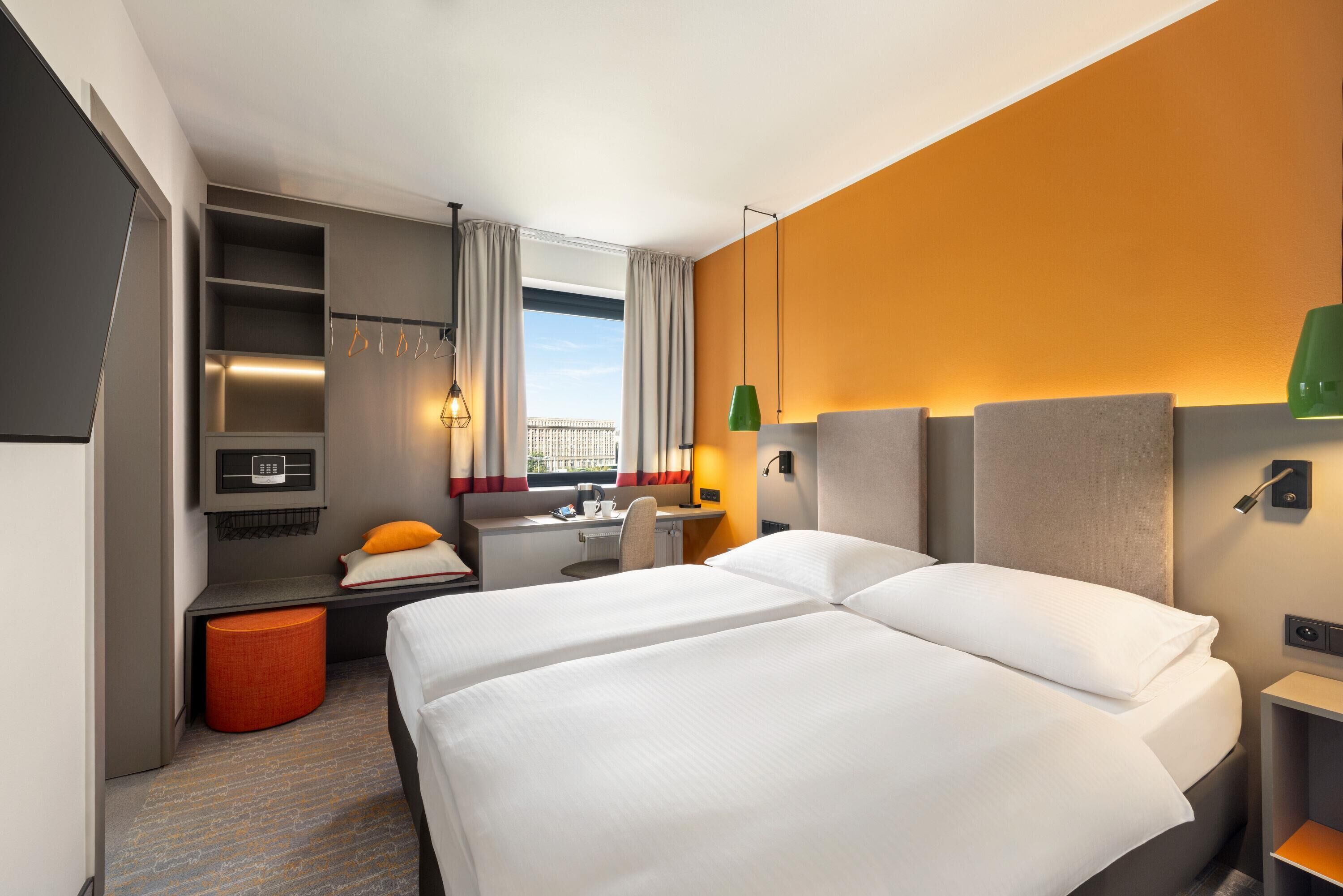 Ett hotellrum med två sängar, ett skrivbord, en stol, en liten orange pall och utsikt över omgivningarna.