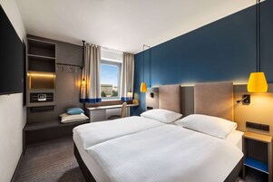 Habitación de hotel con dos camas, un escritorio, una silla, un pequeño rincón de descanso y una ventana con cortinas.