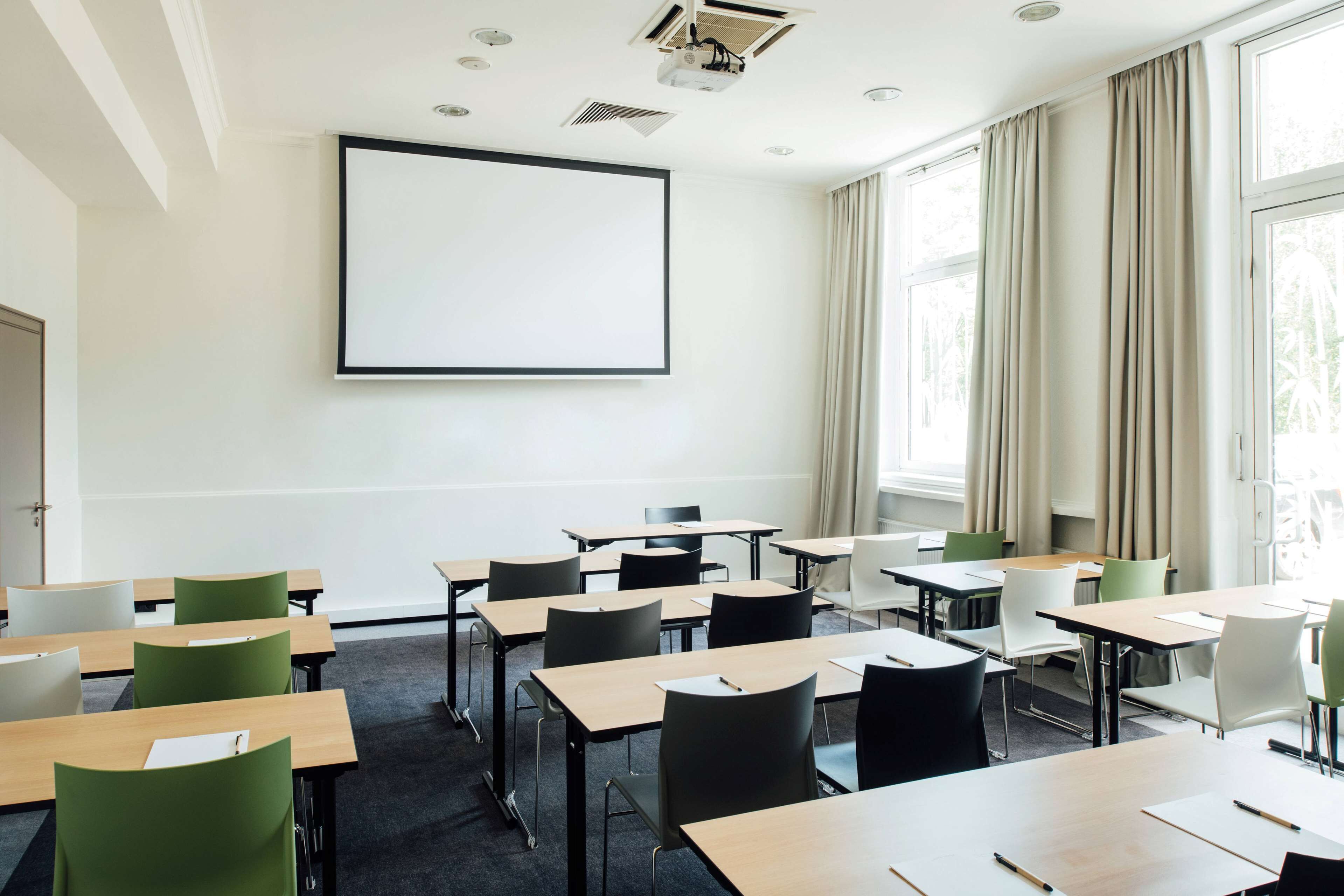 Un aula con escritorios y sillas dispuestas en filas, una pantalla de proyección grande en la pared y ventanas con cortinas.