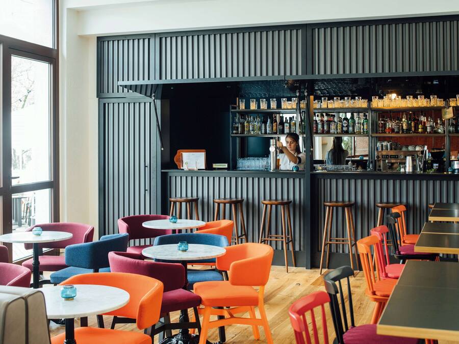 A modern bar area with a variety of seating options including round tables and high stools, a bar counter with a well-stocked shelf of bottles, and a large window providing natural light.
