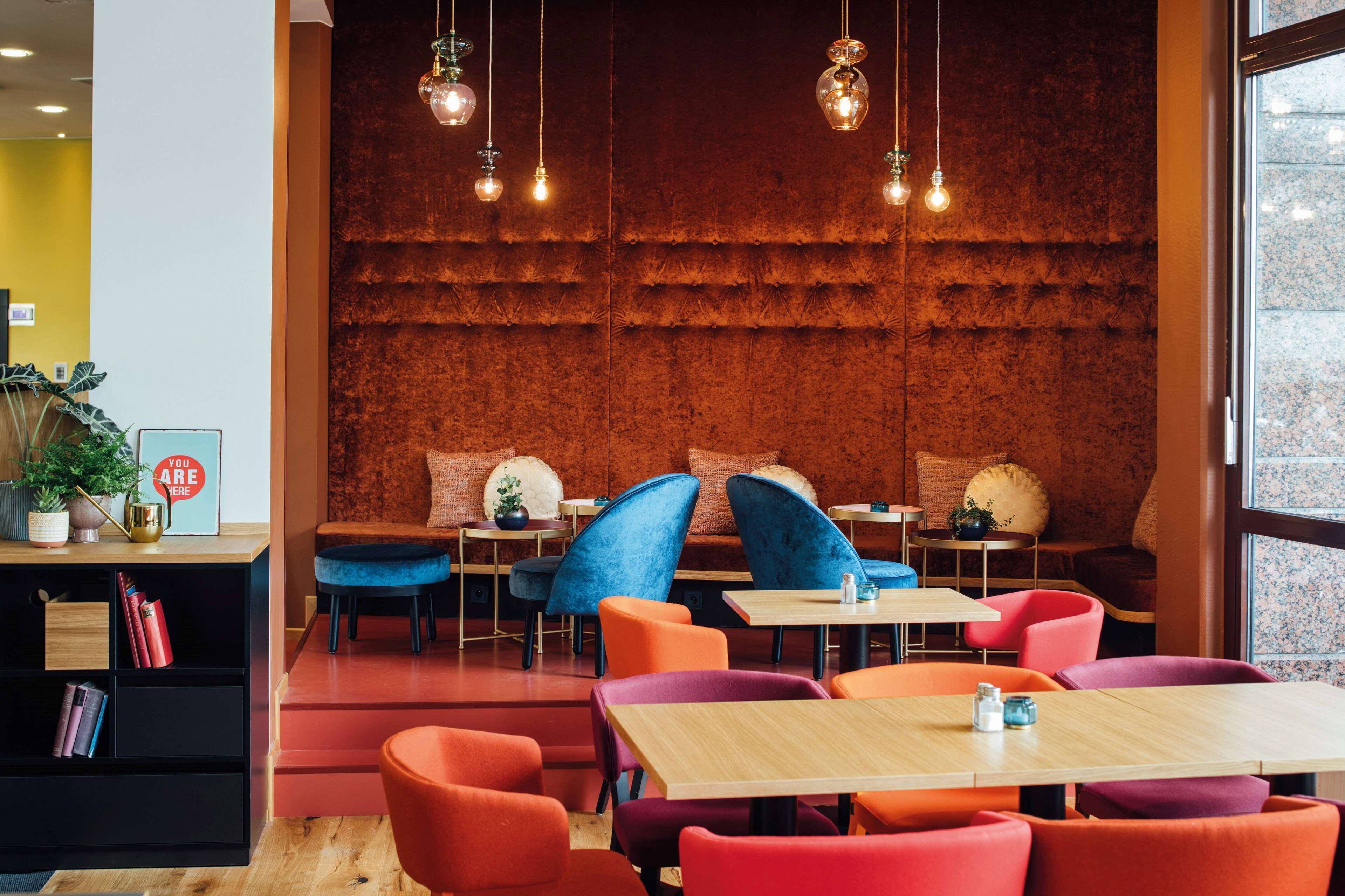 A modern restaurant interior with wooden tables and colorful chairs, a red textured wall, and hanging pendant lights.