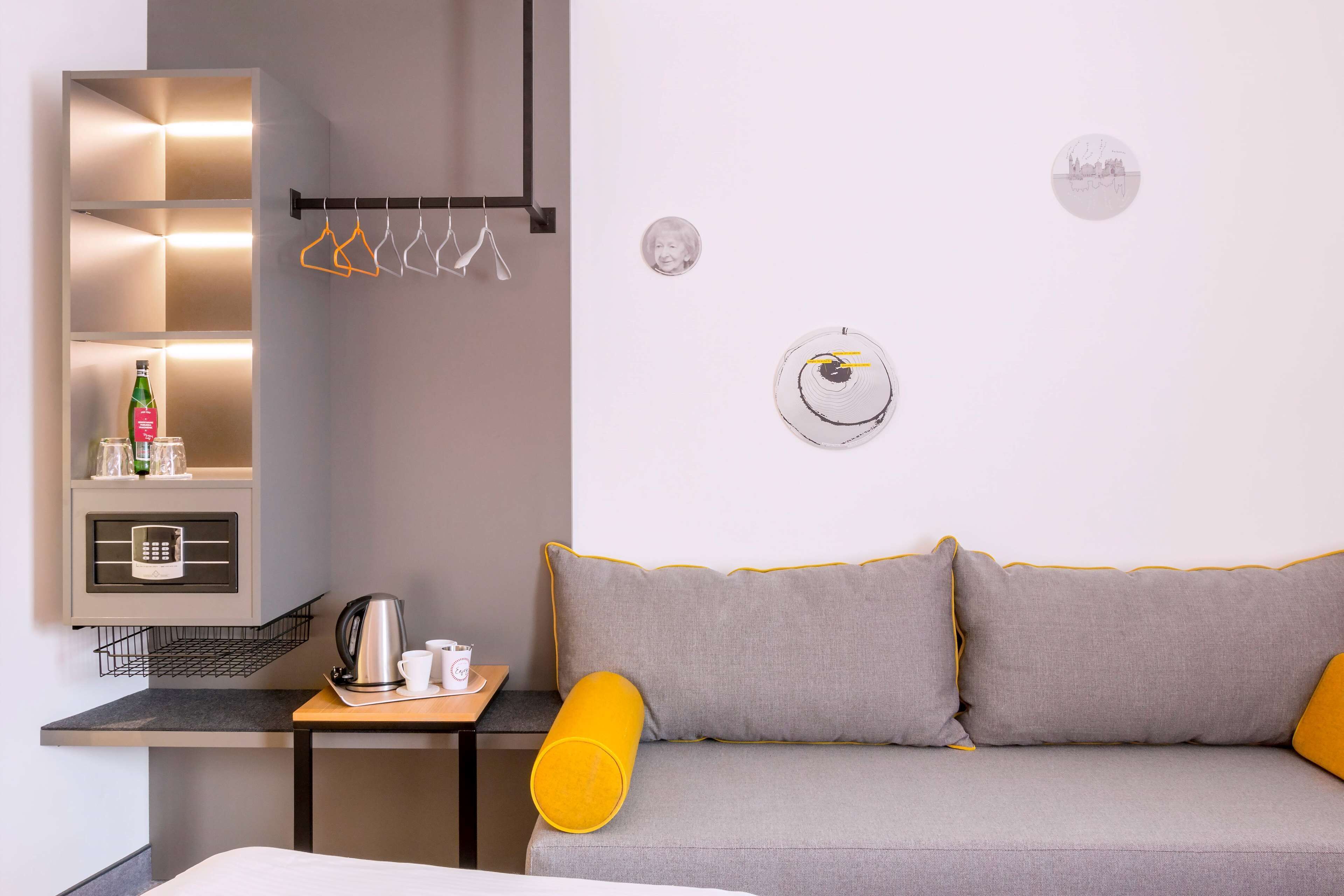 A modern hotel room with a sofa, a small table with a kettle and cups, a shelf with toiletries, and a wall-mounted coat rack.