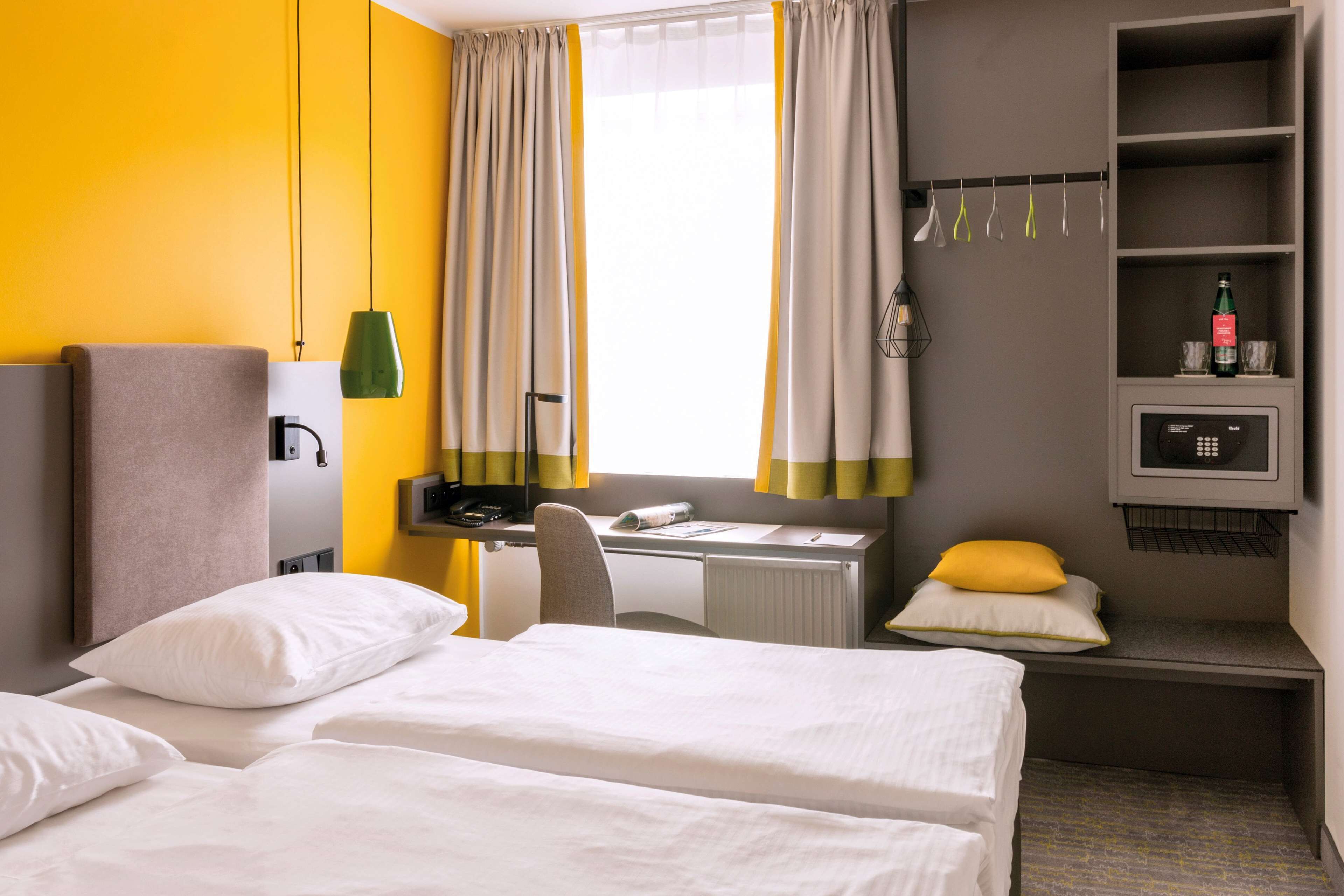 A hotel room with two beds, a desk, a chair, a radiator, a shelf with a bottle, and a window with curtains.