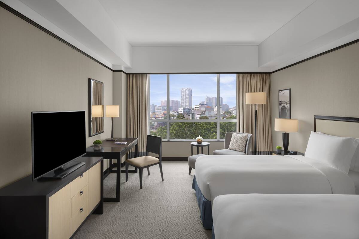 Hotelkamer met een groot bed, een bureau met televisie, een stoel, een klein tafeltje met lamp en uitzicht op de stadshemel.