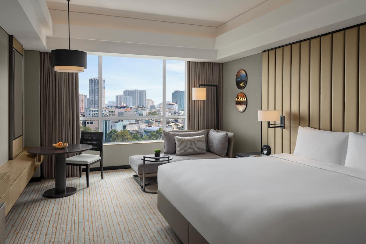 Habitación de hotel con una cama grande, un escritorio con silla, una mesita y vistas de la ciudad a través de amplios ventanales.