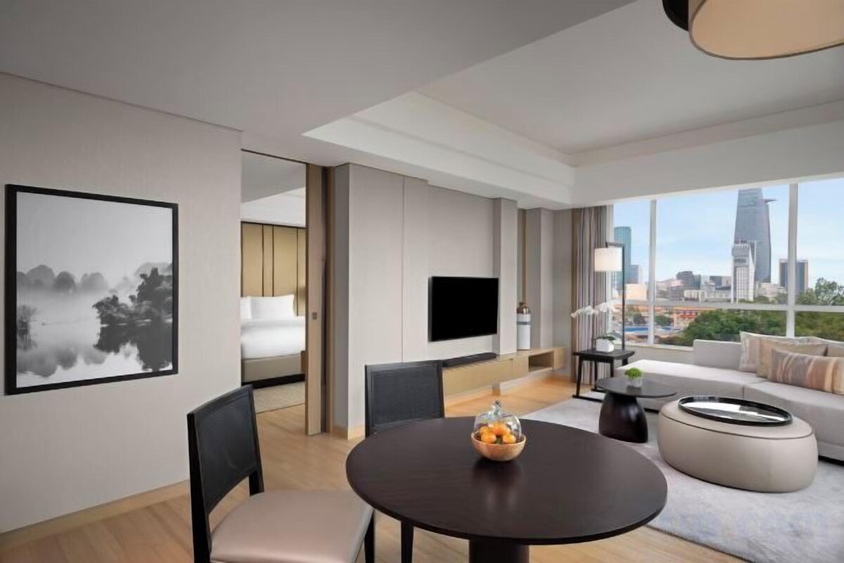 A modern living room with a round dining table, a sofa, and a large window offering a city view.
