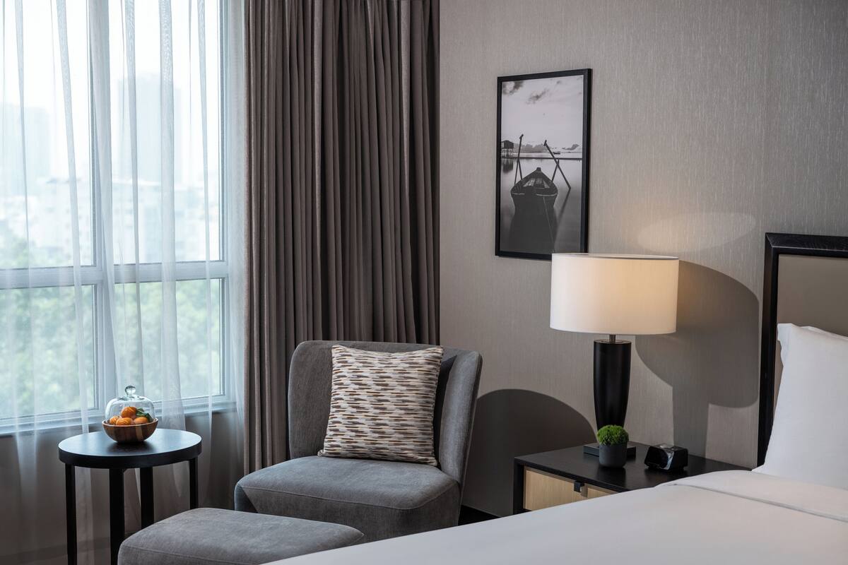Een hotelkamer met een bed, een stoel, een klein tafeltje met een schaal fruit, een lamp en een lijstje aan de muur.