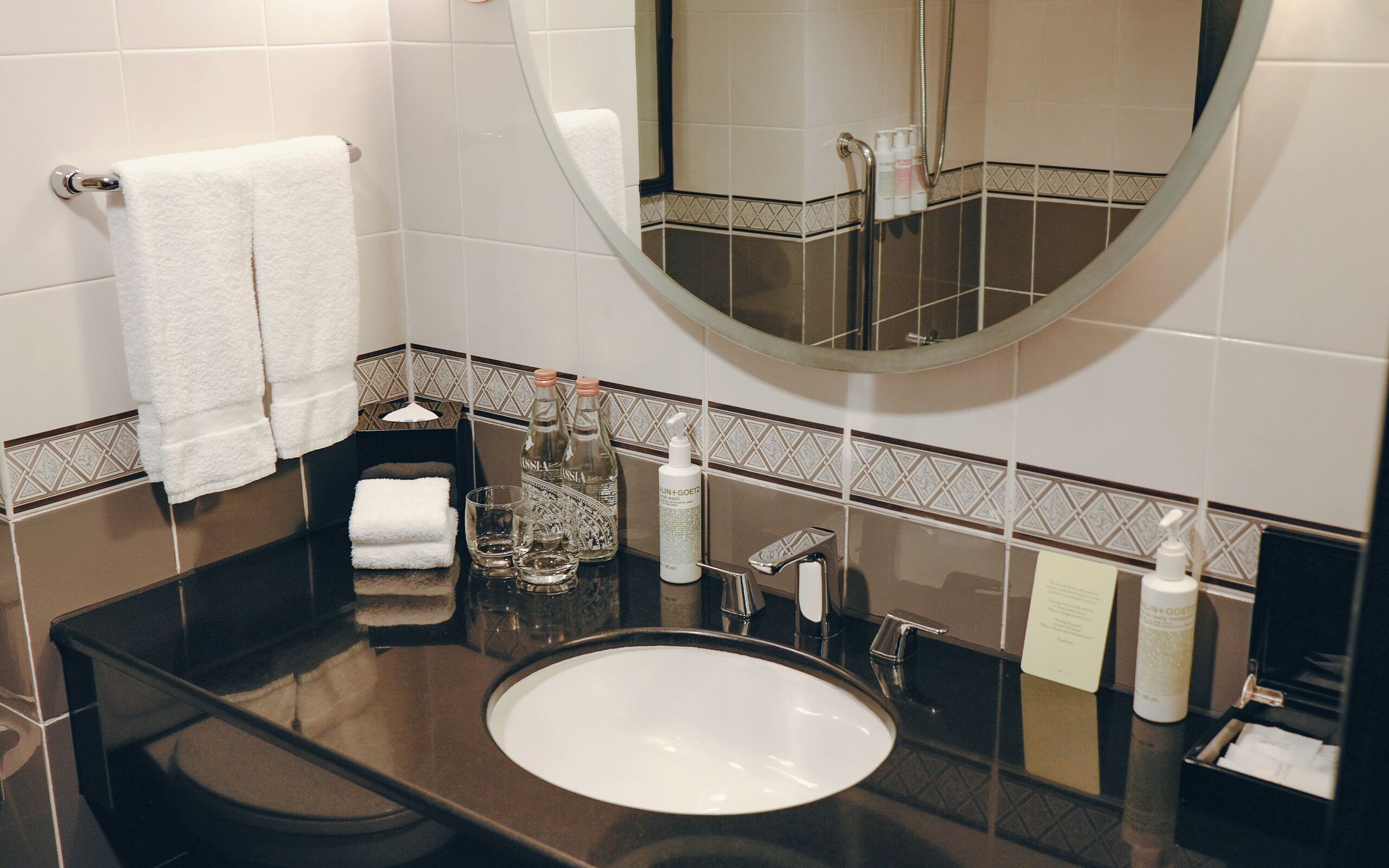Et moderne badeværelse med en sort bordplade, hvid vask og et stort spejl. Der er håndklæder, toiletartikler og et glas på bordet.