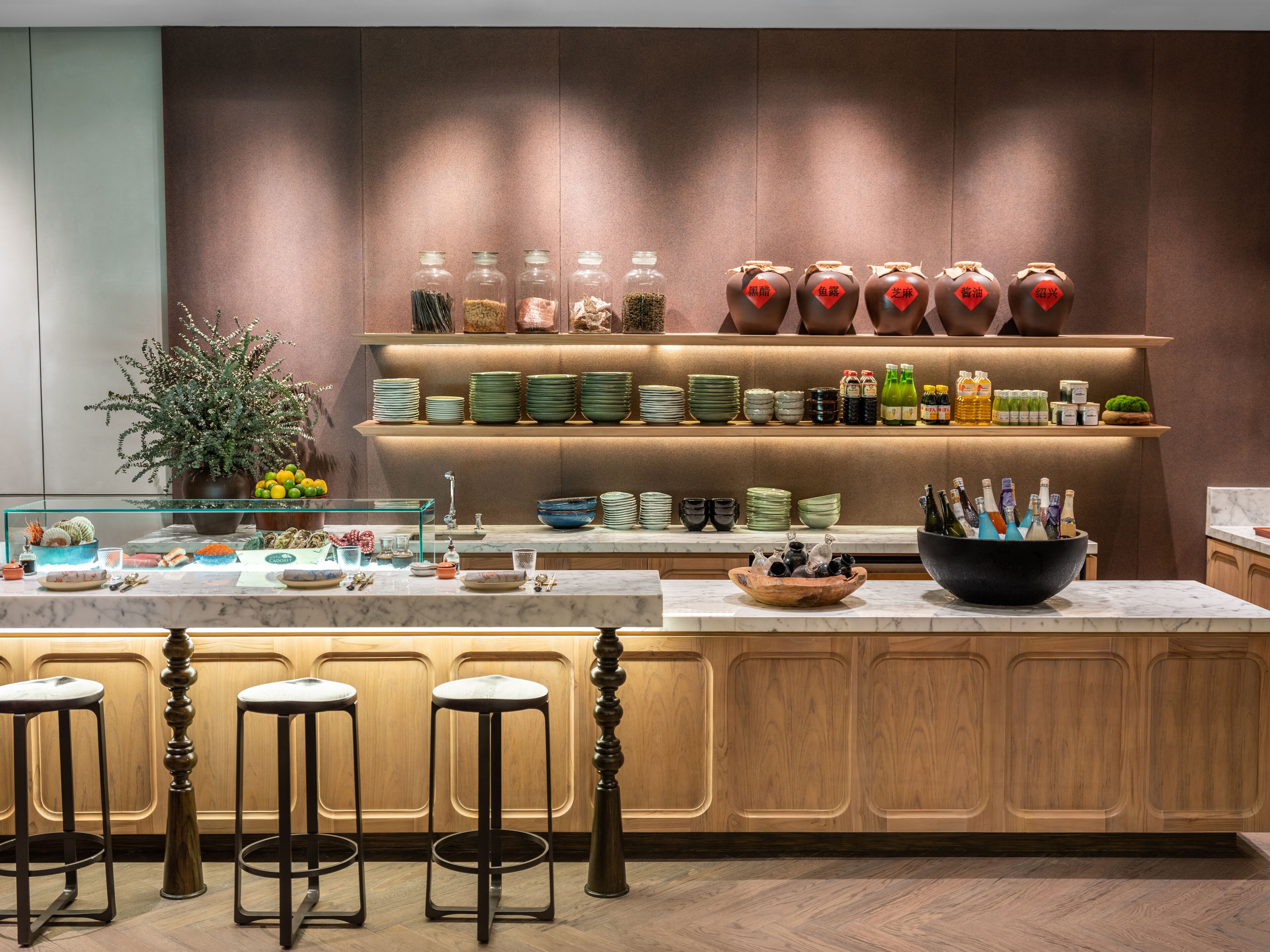 A modern kitchen with a marble countertop, wooden bar stools, and a variety of kitchenware and bottles on shelves.