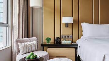 A hotel room with a bed, a nightstand with a lamp, a chair with a cushion, a small table with a bowl of fruit, and a window with curtains.