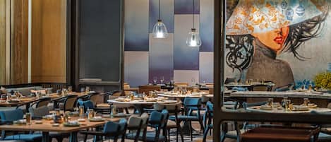 Un restaurante con varias mesas y sillas, un mural grande en la pared e iluminación colgante.
