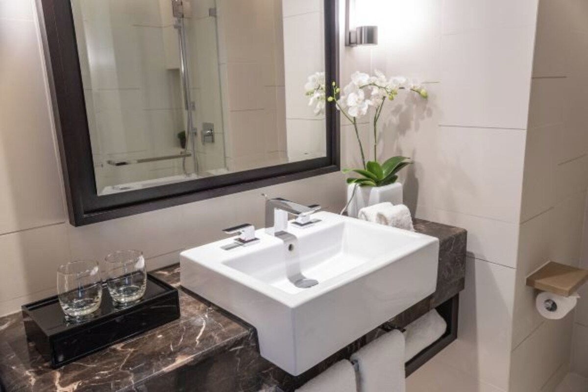 A modern bathroom with a large mirror, a white sink, and a marble countertop. There are two glasses on a tray, a towel rack, and a small shelf with a roll of toilet paper.