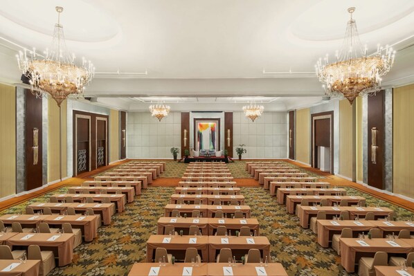 Une salle de conférence avec des rangées de tables, des lustres et un grand écran à l’avant.