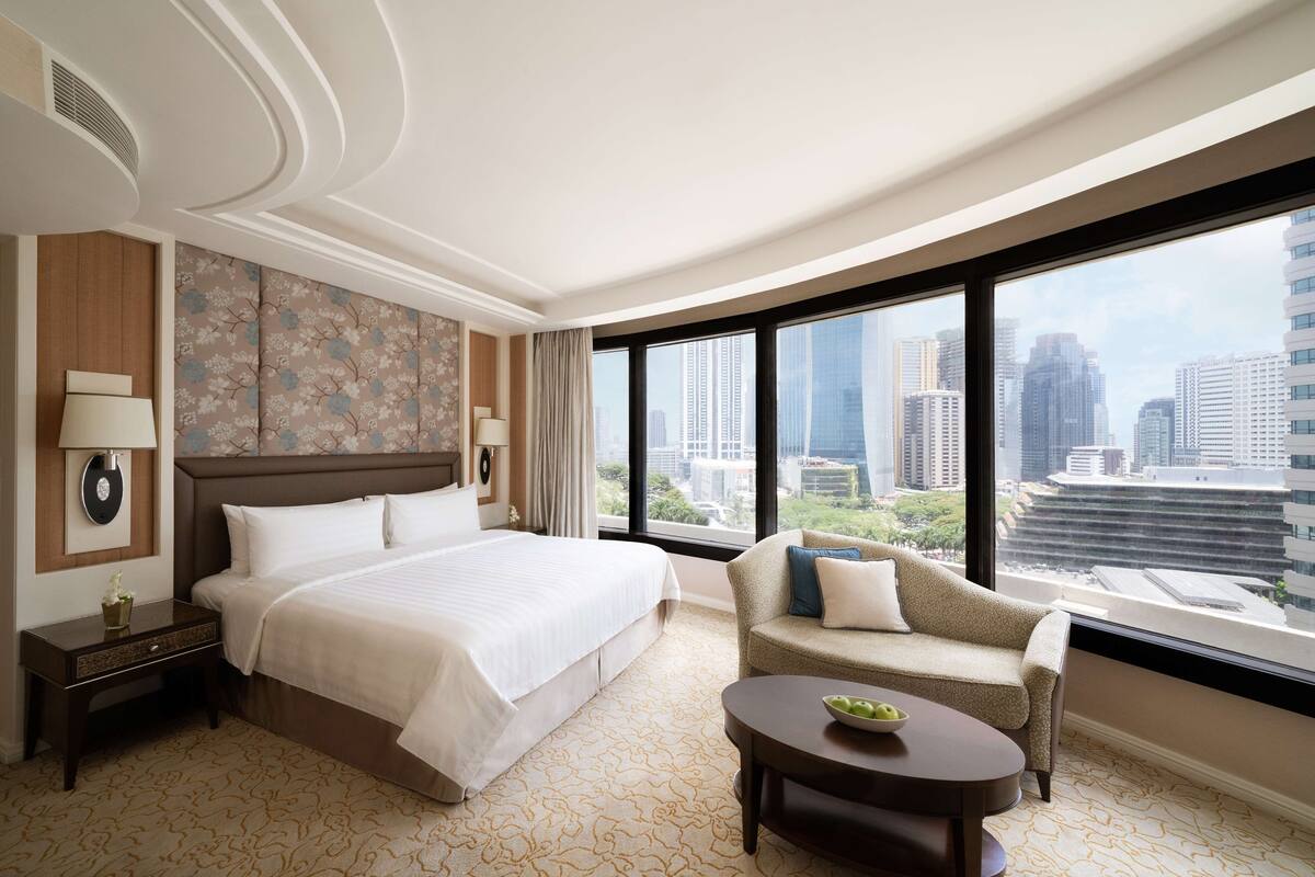 A hotel room with a large bed, a sofa, a small table, and a view of the cityscape through large windows.