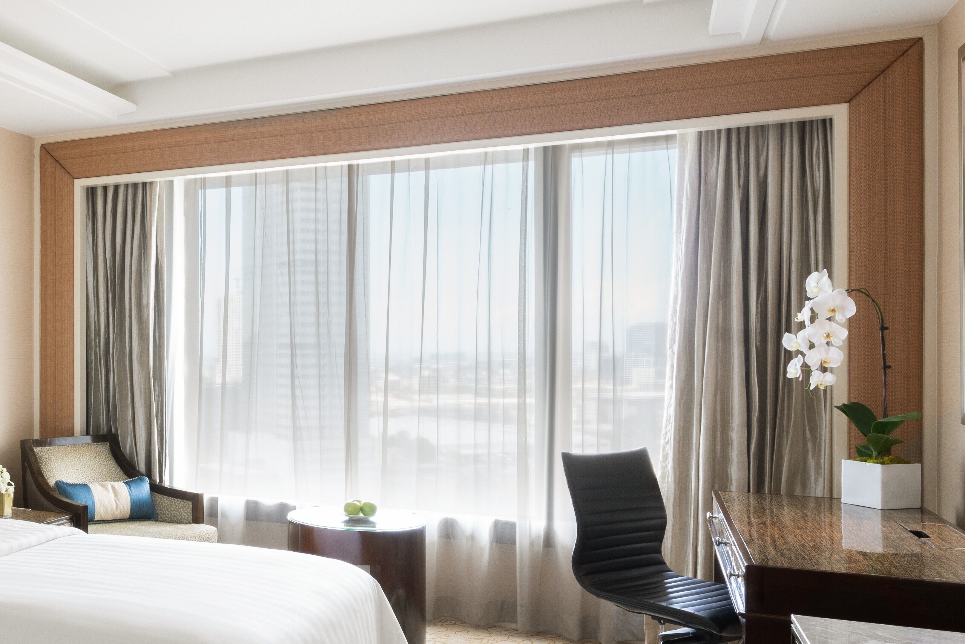 Habitación de hotel con una cama grande, un escritorio, una silla y vistas de la ciudad a través de cortinas translúcidas.