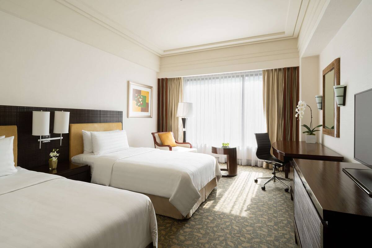 A hotel room with two beds, a desk, a chair, a TV, and a window with curtains.
