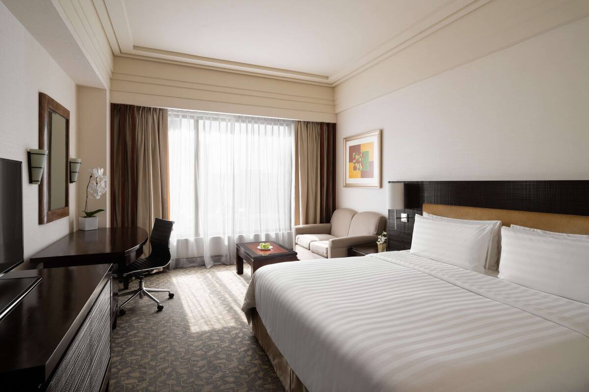 A hotel room with a large bed, a desk with a computer, a chair, a small table with a plant, and a window with curtains.