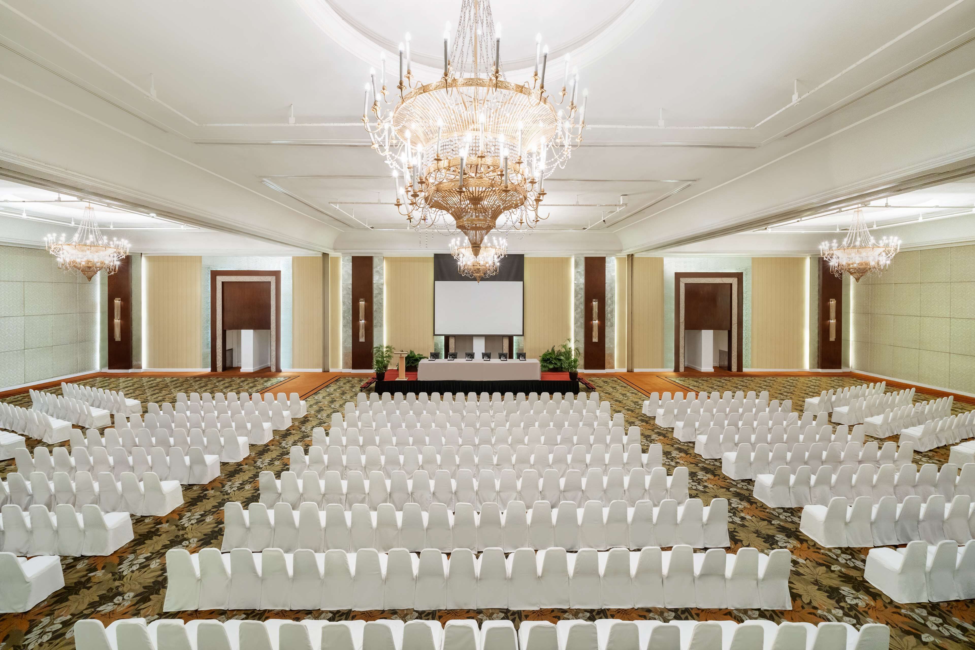 A large conference room with rows of white chairs, a central stage, and a chandelier.