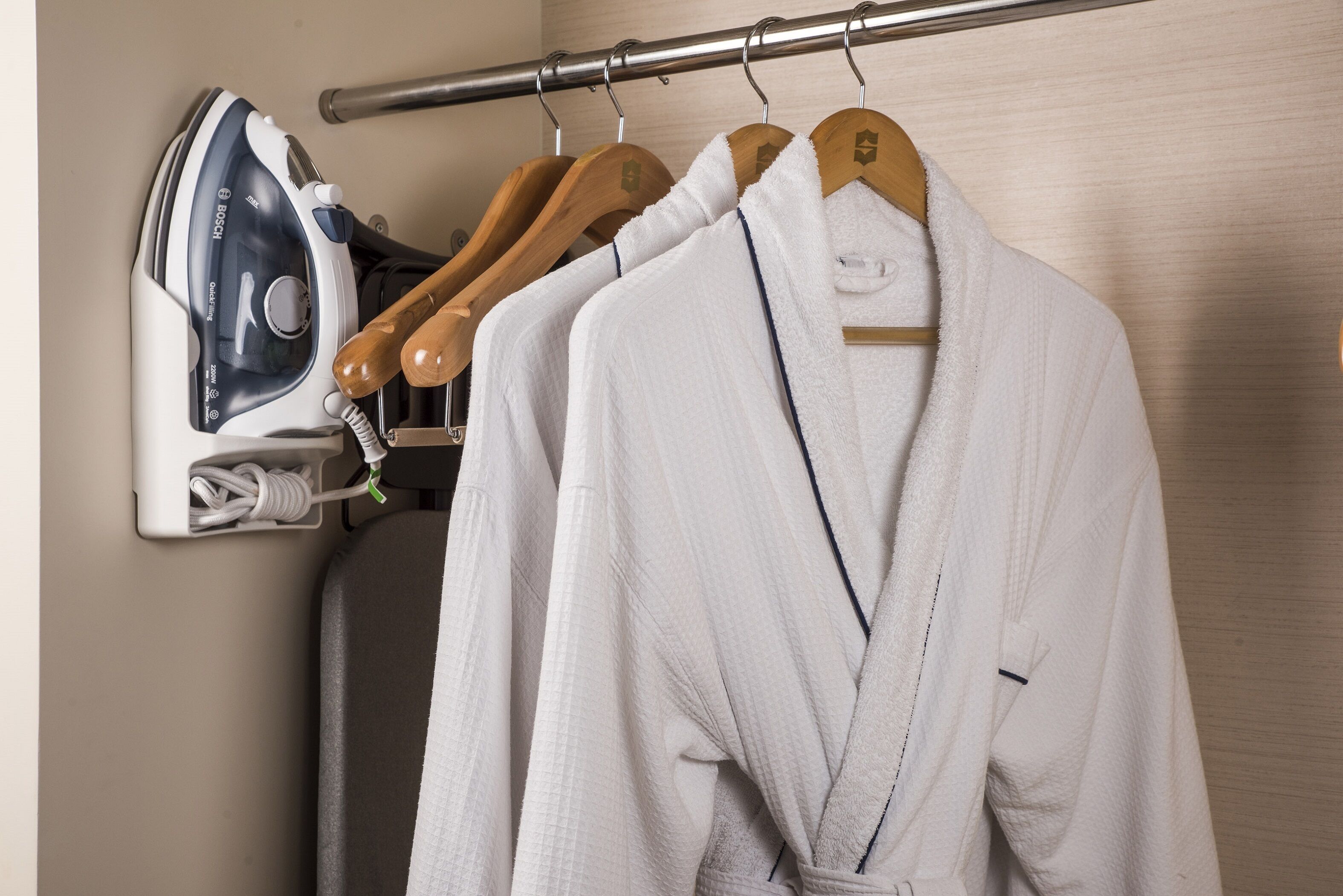 Un baño de hotel con un perchero de madera que sostiene dos batas blancas, un planchador Bosch y un par de zapatos.