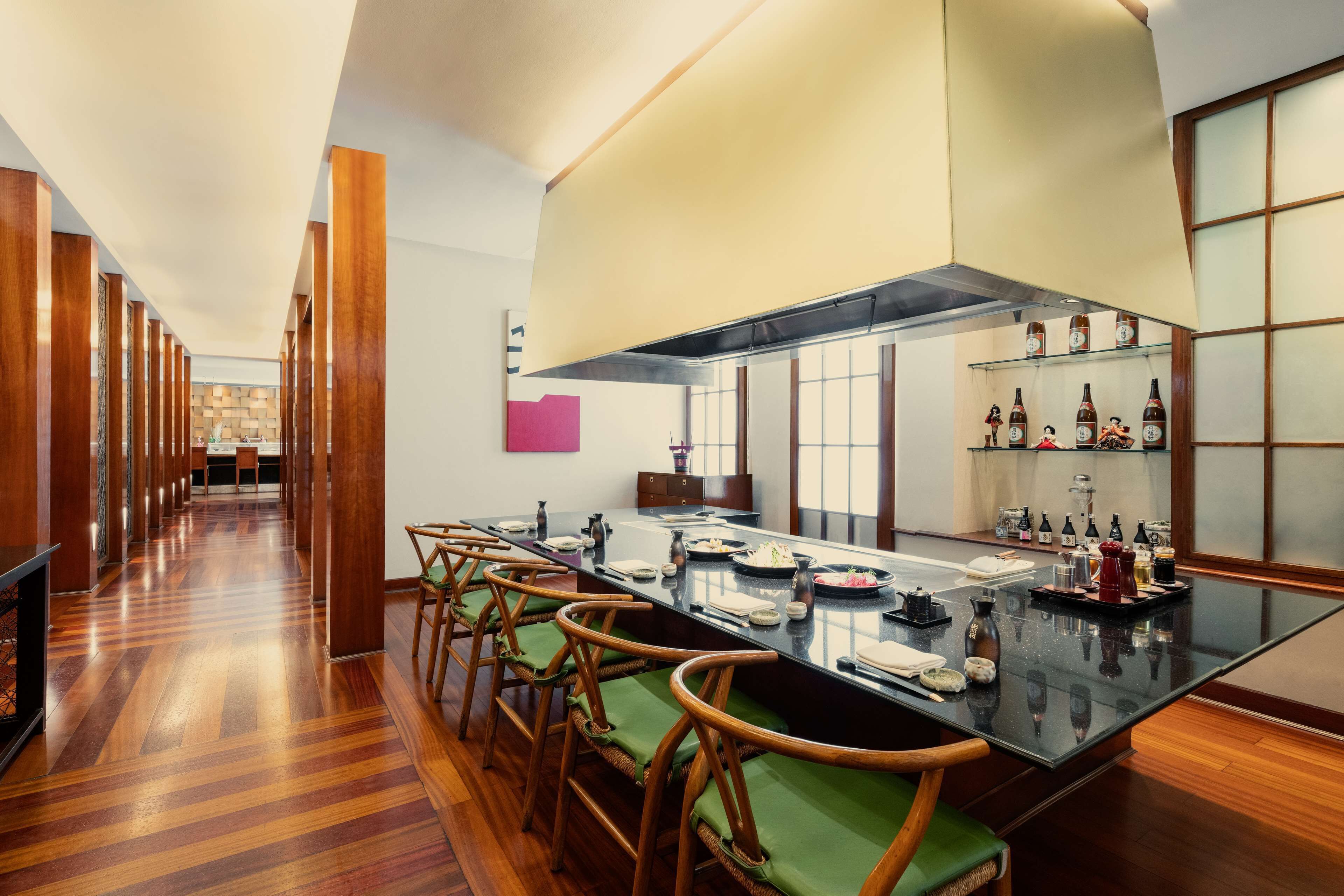 A modern kitchen with a long dining table, wooden chairs, and a variety of bottles on shelves.