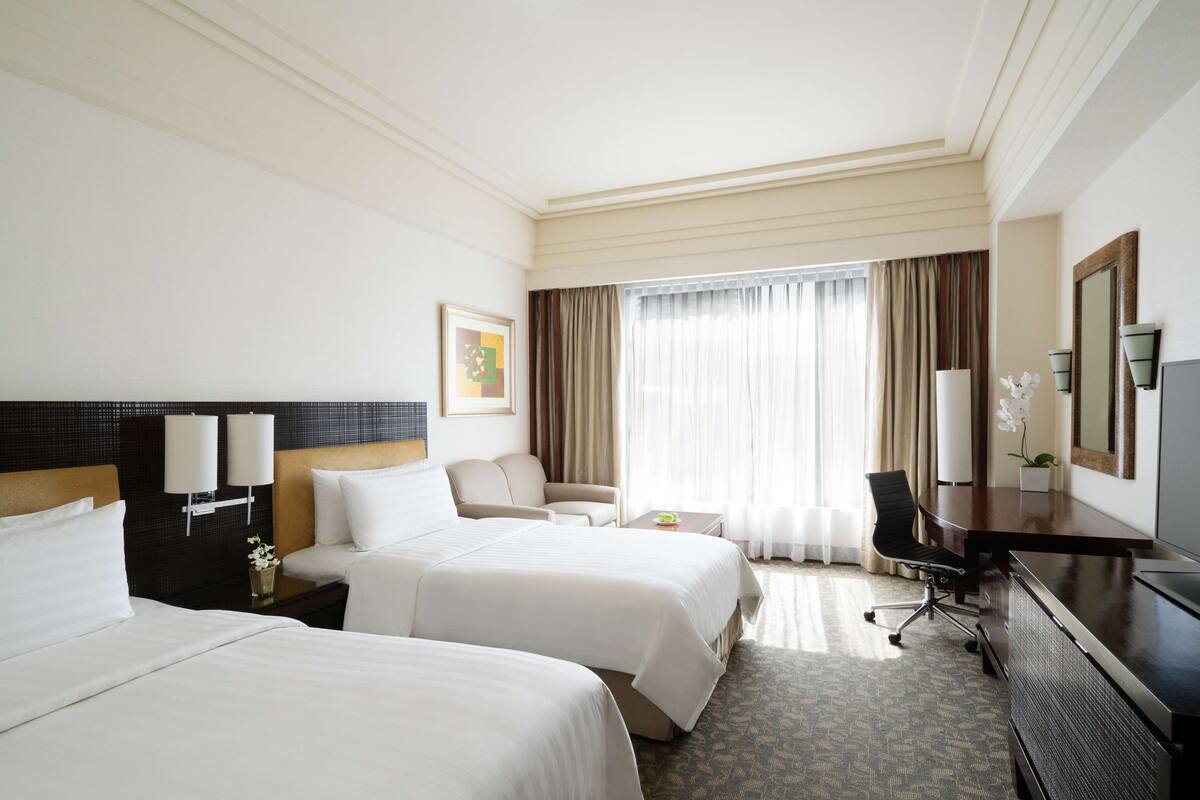A hotel room with two beds, a desk, a chair, a TV, and a large window with curtains.