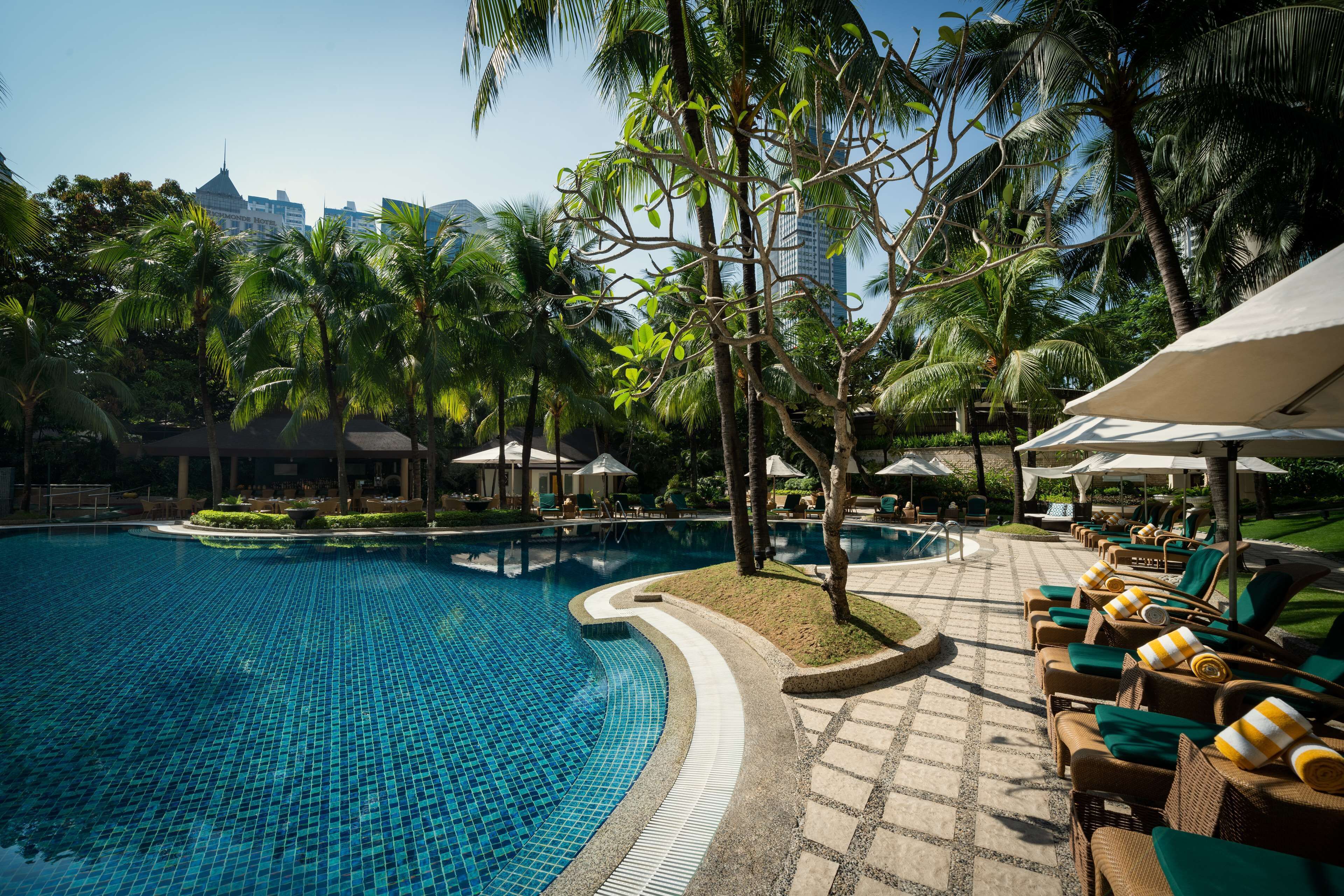 A swimming pool with lounge chairs and umbrellas, surrounded by palm trees and buildings.