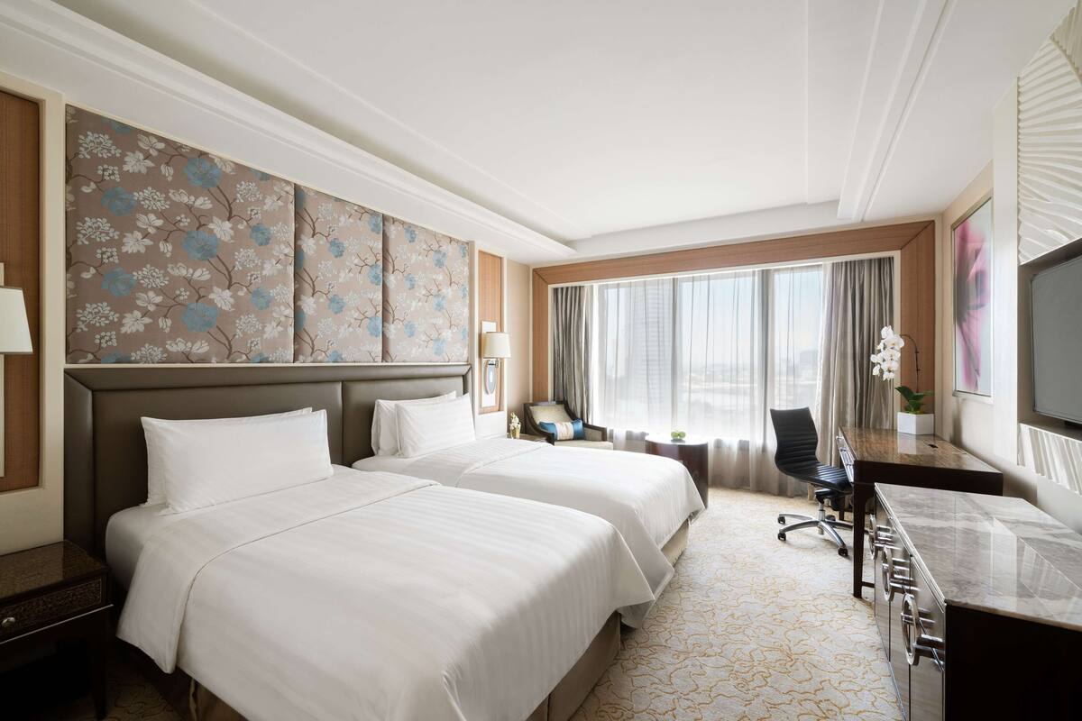 A hotel room with two beds, a desk, a chair, a TV, and a large window with curtains.