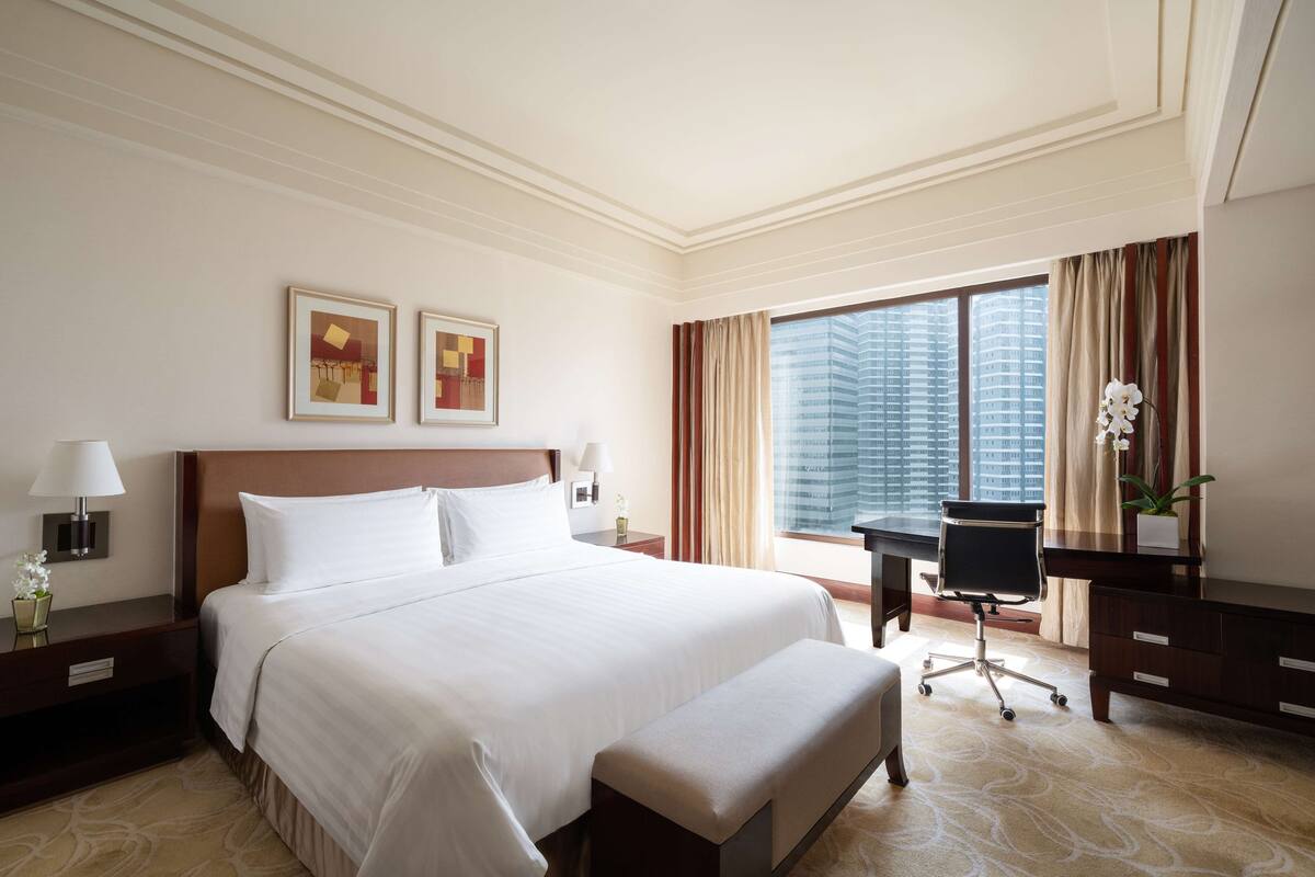 A hotel room with a large bed, a desk, a chair, a nightstand, and a window with a view of buildings.