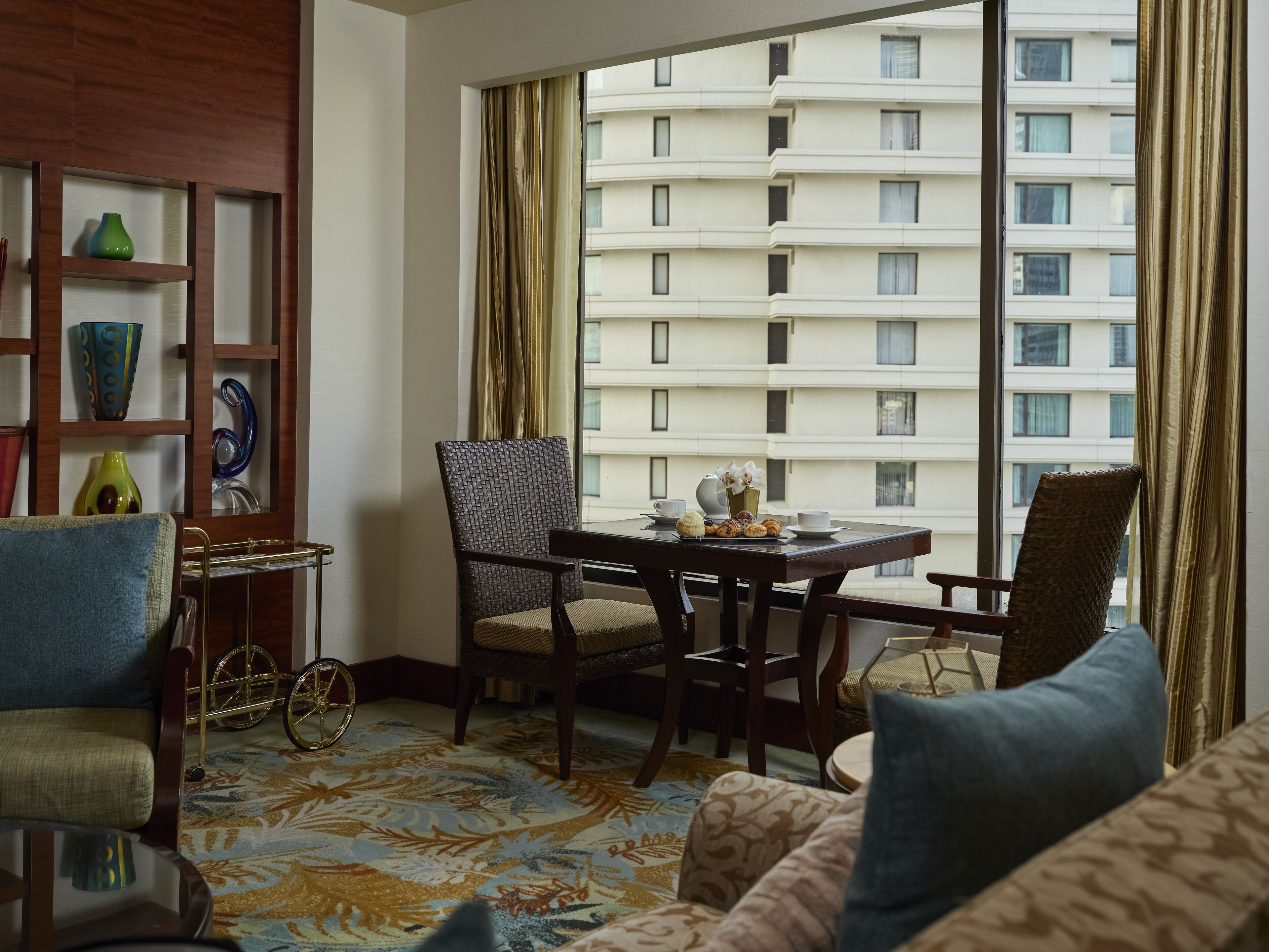 A hotel room with a large window, a dining table with chairs, a sofa, a bookshelf, and a decorative bicycle wheel.