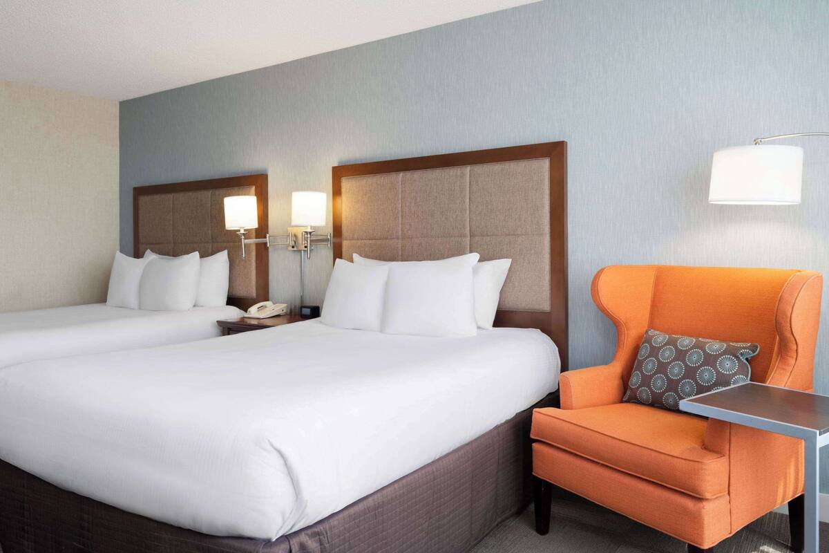 A hotel room with two beds, a nightstand with a phone, an orange armchair, and a small table.