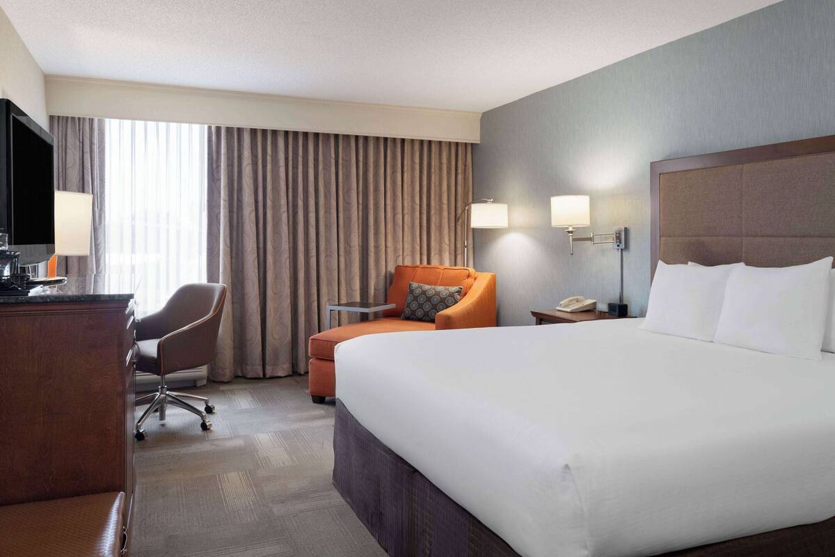 A hotel room with a large bed, a desk with a lamp, a chair, and a TV.