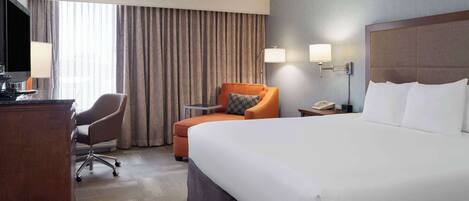 A hotel room with a large bed, a desk with a lamp, a chair, and a TV.