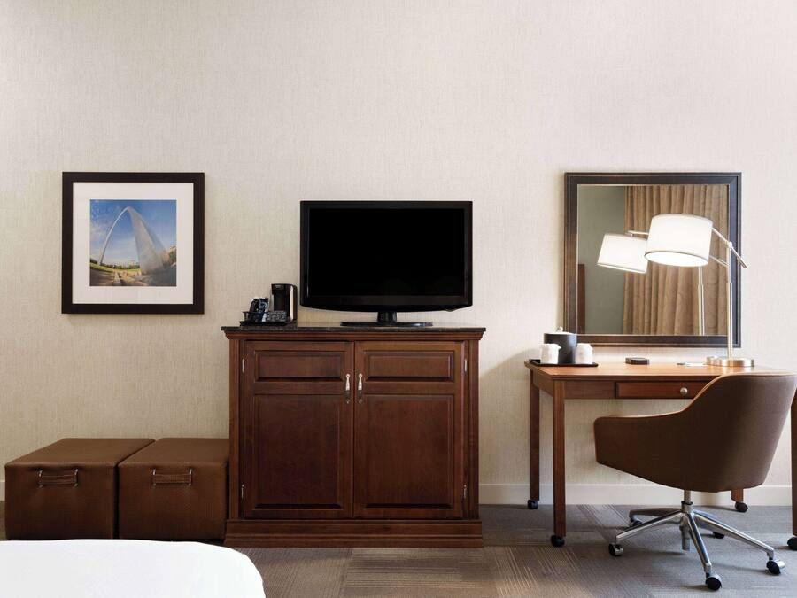 Quarto de hotel com cama, armário de madeira com TV de tela plana, escrivaninha com cadeira, abajur e quadro na parede.