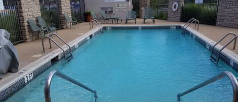 A rectangular pool with a tiled edge and metal handrails. There are chairs and a lifeguard chair near the pool. A sign on the wall indicates no diving and no lifeguard on duty.