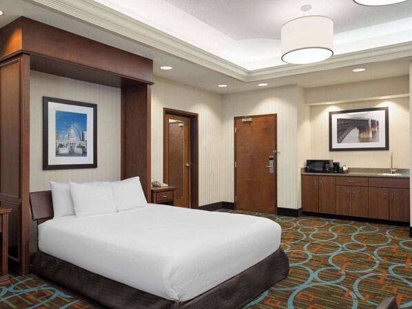A hotel room with a bed, a desk, a mirror, and a framed picture of a bridge. - Wingate by Wyndham St. Louis Airport (St. Ann)