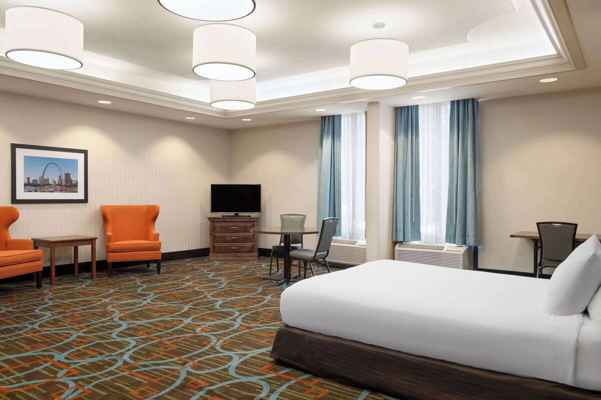 A hotel room with a large bed, two armchairs, a TV, a small table, and a window with curtains.