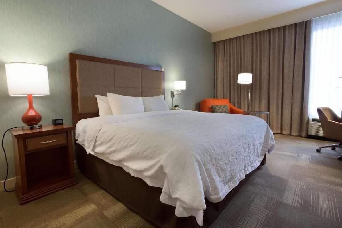 A hotel room with a large bed, a nightstand with a lamp, a chair, and a window with curtains.