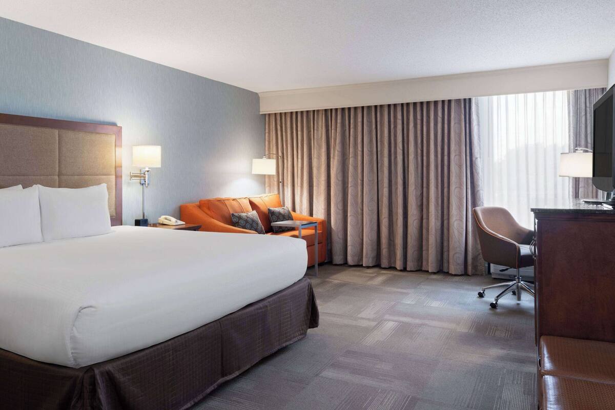 A hotel room with a large bed, a desk, a chair, and a view of the outside through curtains.