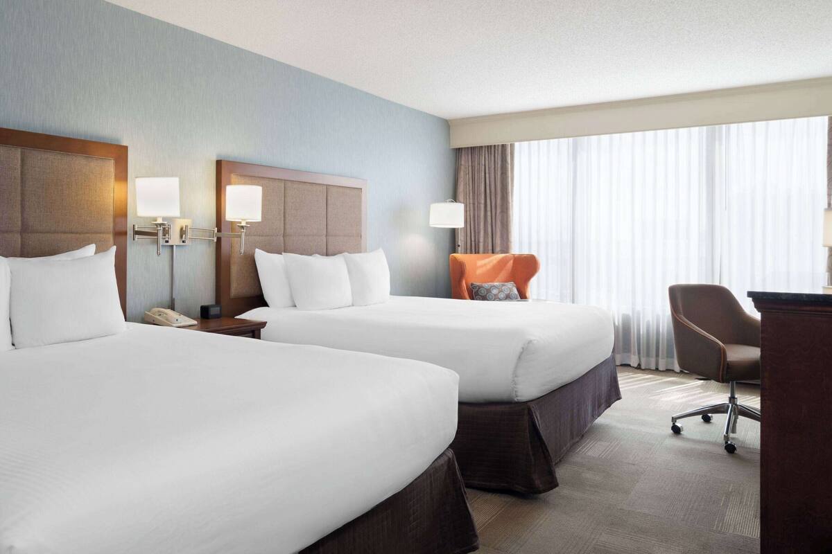 A hotel room with two beds, a desk, a chair, and a large window with curtains.