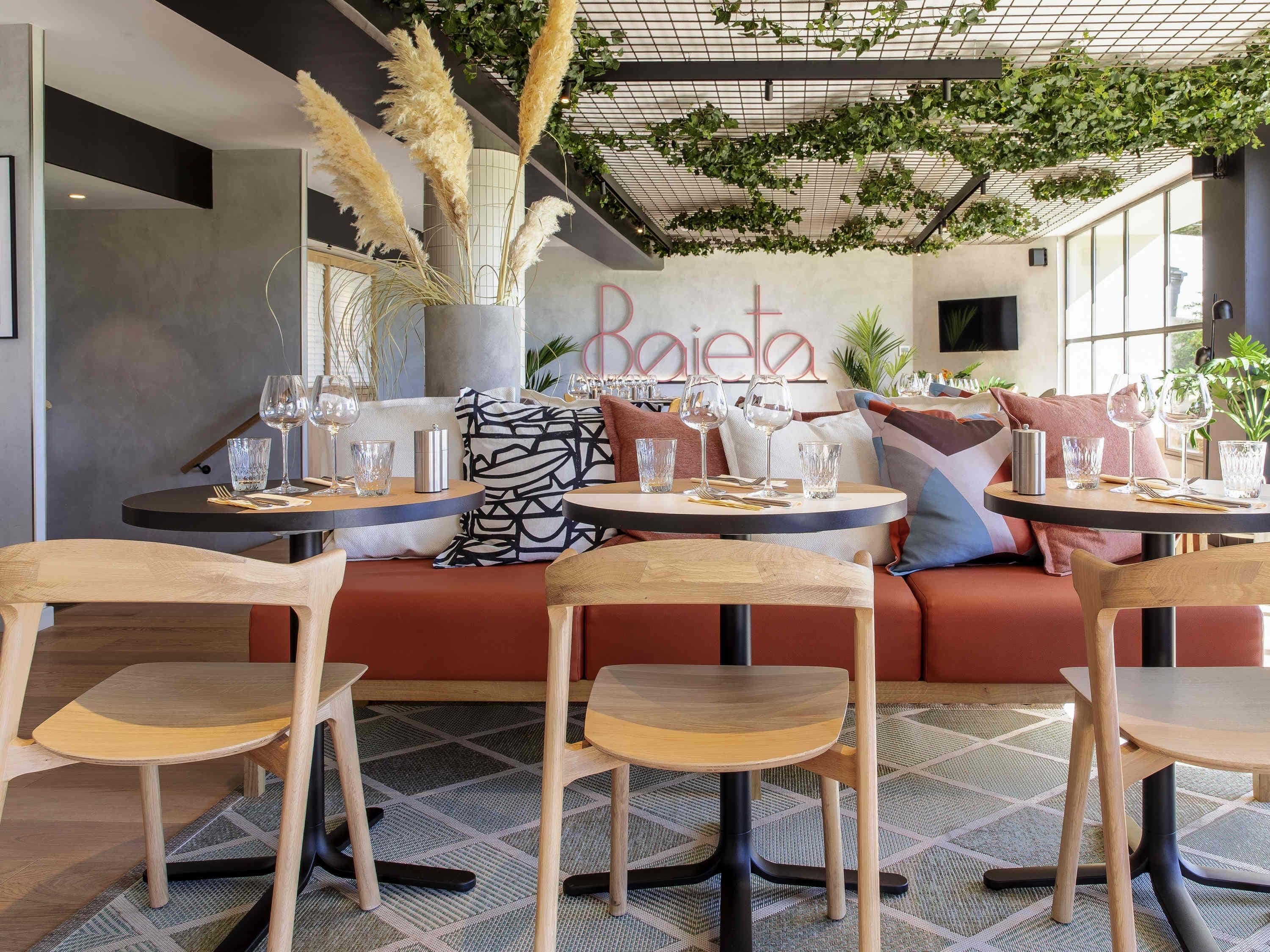 A modern restaurant interior with round tables, wooden chairs, and a cozy seating area with red cushions.