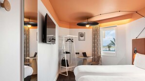 A hotel room with a bed, a desk, a chair, a wardrobe, a mirror, a TV, and a window with a view. - ibis Nice Aéroport Promenade des Anglais (Nice)